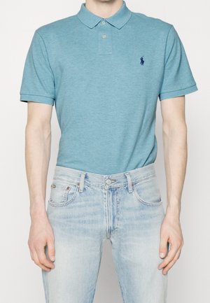 Homme portant un polo ajusté à manches courtes couleur sarcelle et un jean bleu clair, debout devant un fond uni.