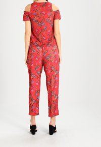 Röd blommig byxdress med kall axel-design, med korta ärmar och cropped-byxor med en svalkande textur och svarta accent-skor.