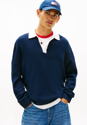 Młody mężczyzna ubrany w granatowy sweter na białej koszuli z kołnierzykiem i czerwonym t-shircie, niebieskie dżinsy oraz niebieską czapkę z prostokątnym logo.
