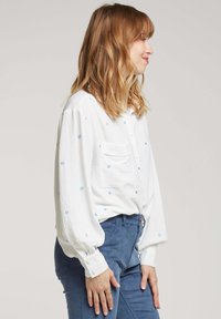 Chemise blanche à manches longues avec broderie florale bleue, poignets à boutons et une poche poitrine, associée à un pantalon en velours côtelé bleu. Tissu texturé.