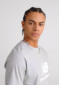 Pilkas ilgomis rankovėmis marškinėliai su baltu logotipu, tekstūruotas audinys, apvalus kaklas. Modelis dėvi sidabrinę grandinėlę, plaukai surišti į kasas. Paprasta fono nuotrauka.