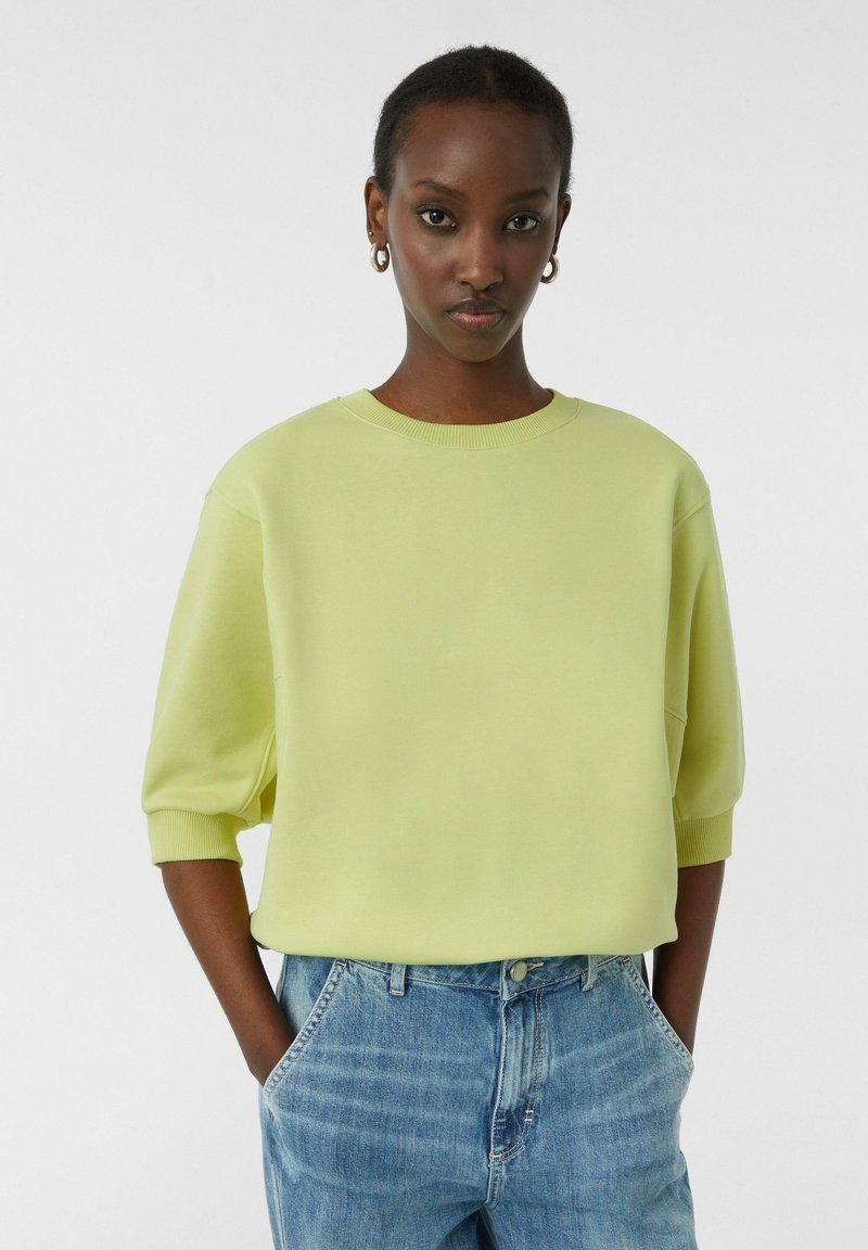 Jasnożółty, oversize'owy sweter z krótkim rękawem i prążkowanym dekoltem, zestawiony z wysokim stanem, wyblakłymi niebieskimi dżinsami.