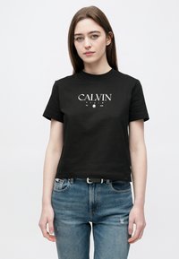 Tricou negru din bumbac cu inscripția "CALVIN KLEIN" imprimată alb, mâneci scurte, decolteu rotund și croială dreaptă. Purtat cu blugi denim albaștri.