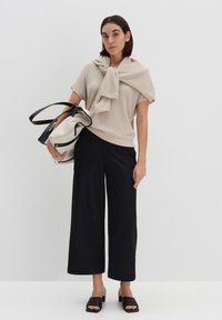 Beige ribbeltricot top met knoopdetail, zwarte wijde broek en zwarte instapsandalen. Draagt een grote, gestructureerde zwart-witte tas.