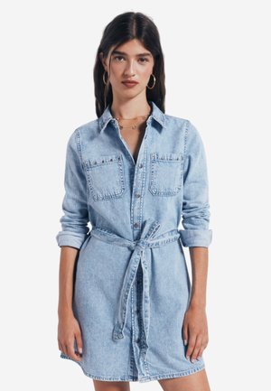 Robe chemise en denim bleu clair avec des manches longues, un devant boutonné, deux poches poitrine et une taille nouée. Présente un col classique et des poignets retroussés.