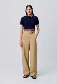Maglione cropped blu navy sovrapposto a una camicia a quadri con colletto, abbinato a pantaloni cargo beige larghi con tasche laterali, indossato con sandali neri.