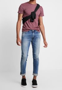 Merlot-Flaum Crewneck-T-Shirt, schwarzer Strickpullover über dem Körper drapiert, hellblaue, distressed Jeans, schwarze Sneakers, hochgekrempelte Säume.