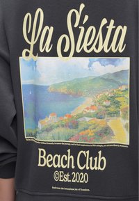 Σκούρα φούτερ με μεγάλο γραφικό στο μπροστινό μέρος. Απεικονίζει μια παραθαλάσσια τοποθεσία με κτίρια, λουλούδια και γαλάζιο ουρανό. Το κείμενο αναγράφει «La Siesta Beach Club Est. 2020.»