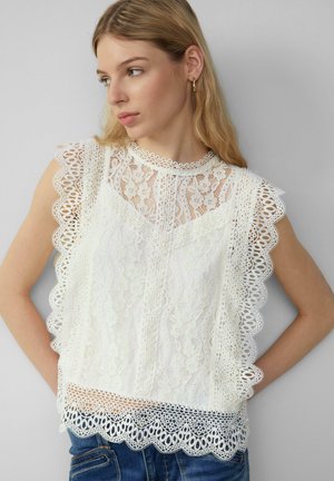 Weißes Spitzenoberteil mit gewellten Kanten, floralen Mustern und hohem Kragen. Kombiniert mit Jeansshorts für einen Kontrast.