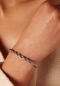 Zilveren gedraaide armband om een pols, met een gladde, glanzende textuur en een gevlochten ontwerp. De achtergrond is neutraal en zacht.