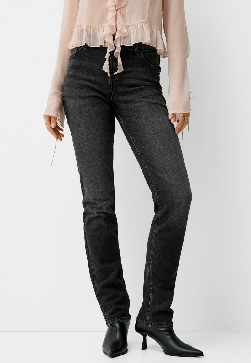 Donna con jeans grigi scuri aderenti, stivali neri con tacco a punta e una blusa leggera rosa con volant, maniche lunghe e anelli alle dita.