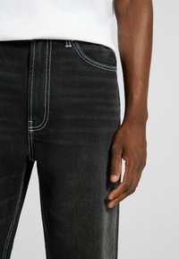Jeans in denim nero con cuciture bianche a contrasto, caratterizzati da un design a cinque tasche e un tessuto testurizzato, mostrati accanto a una mano.