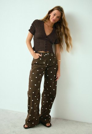 Frau mit langen Haaren, die ein strukturiertes braunes Kurzarm-Oberteil und braune, weit geschnittene Hosen mit weißen Punkten trägt, steht vor einer hellen Wand.