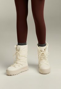 Białe ocieplane zimowe buty ze wysoką cholewką, sznurowane, z gumową podeszwą oraz teksturowanymi akcentami przy kostce i na palcach.