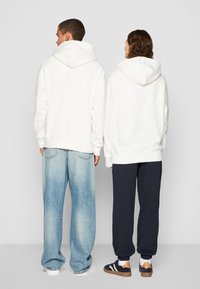Hvite hettegensere sett bakfra, med kengurulomme. En person har på seg falmede blå jeans, den andre mørke joggebukser.