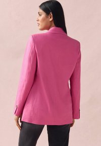 Růžový blazer z lnu, má přiléhavý střih, špičaté klopy a knoflíkové manžety. Nosený s tmavými kalhotami na světlém pozadí.