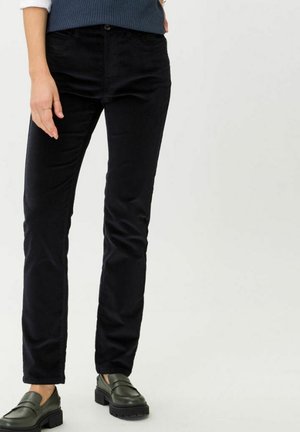 Person trägt eng anliegende schwarze Hose und dunkelgrüne Loafers mit dicker Sohle, steht vor einem einfarbigen hellen Hintergrund.