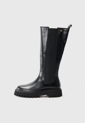 Stivale in pelle nera al ginocchio, con suola spessa a denti, pannello elastico ai lati e linguetta sul retro, su sfondo neutro.