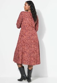 Robe longue dans un motif abstrait rouille et orange. Dotée de longues manches, d'une taille cintrée et d'une jupe plissée. Associée à des bottes de cowboy noires.