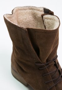 Bottine en daim marron avec une surface texturée, fermeture à lacets à l'avant et doublure douce. Dotée d'un talon en bois empilé et d'un bord brut en haut.