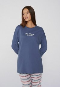 Top de manga larga azul marino con el texto "No more drama" en blanco. Combinado con pantalones de pijama de chevron multicolor y estampado. Tejido suave, corte relajado.