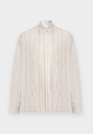 Homme aux cheveux bouclés portant une chemise à manches longues beige à rayures, un pantalon large beige clair et des chaussures noires, debout avec une main dans la poche.