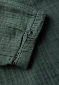 Tessuto verde scuro con una superficie texturizzata e spiegazzata. Presenta un polsino arricciato con cuciture visibili, creando un aspetto morbido e casual.