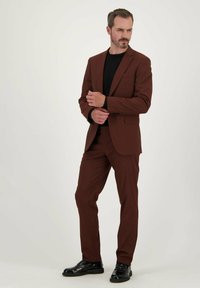 Bruin op maat gemaakt pak met een gestructureerde stof, slim fit, met een notch-lapel, en een zwart T-shirt met ronde hals. Glanzend zwarte dresschoenen maken de look compleet.