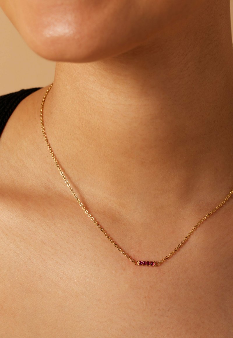 Gouden ketting met een delicaat barpendant waarop vijf rode edelstenen zijn geplaatst, met een gladde textuur en eenvoudig ontwerp.