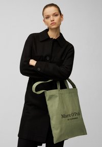 Femme en manteau noir tenant un sac fourre-tout vert olive avec le texte "Marc O'Polo EST. À STOCKHOLM" sur le devant, sur fond uni.
