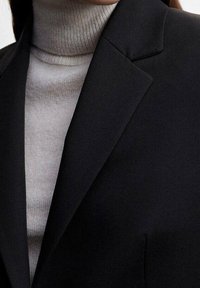 Musta bleiseri, jossa on rakenteelliset kaulukset harmaan poolopaidan päällä. Siloinen kankaan rakenne näkyy, esittäen puhtaita linjoja ja vartalonmyötäistä istuvuutta.