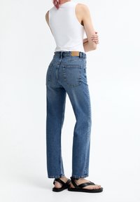 Džíny s vysokým pasem, modré džíny s rovnými nohavicemi, s kapsami na zadní straně a hnědou koženou nášivkou na pase. Nosené s černými sandály.