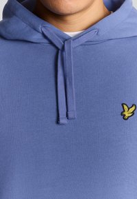Blå hættepullover med struktureret stof, der har et lille gult broderet logo på brystet og snore ved halsåbningen.