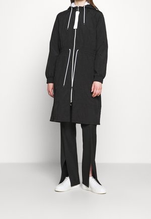 Personne portant une longue veste noire à capuche avec des cordons blancs, un pantalon noir avec des fentes à l'avant et des baskets blanches, debout sur un fond uni.