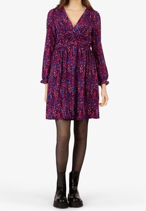 Donna che indossa un vestito a maniche lunghe, lungo fino al ginocchio, con motivi viola e rosa, calze nere e stivaletti neri, in piedi su uno sfondo bianco.