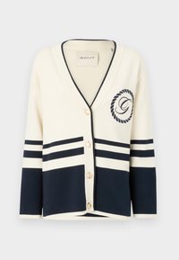 Cardigan crem cu dungi bleumarin, decolteu în V, cinci nasturi aurii și detaliu brodat cu logo. Fabricat dintr-un material moale, texturat.