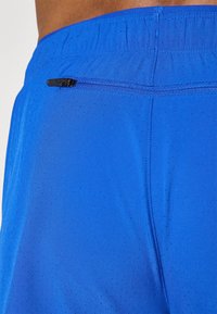 Calças de desporto azuis com uma textura respirável e suave, apresentando perfurações. Inclui um bolso com fecho nas costas para armazenamento seguro.