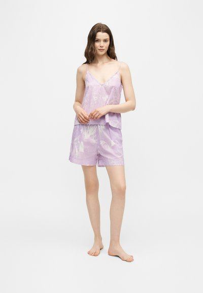 Jeune femme debout pieds nus portant un haut sans manches à fleurs violet clair et un short assorti sur un fond uni.