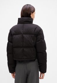 Casaco puffer preto com gola alta, apresentado num design largo e com costura matelassada. Comprimento curto com uma superfície suave e texturizada.