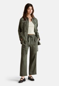 Chaqueta de lino verde oliva con cintura ajustable, combinada con pantalones de cintura elástica a juego y zapatos negros tejidos, llevada sobre una blusa clara.