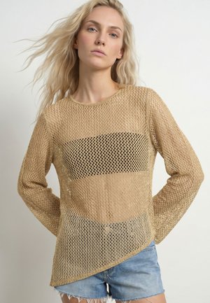 Femme aux longs cheveux blonds portant un pull en maille beige ajourée sur un top noir et un short en denim bleu clair, debout les mains dans le dos.