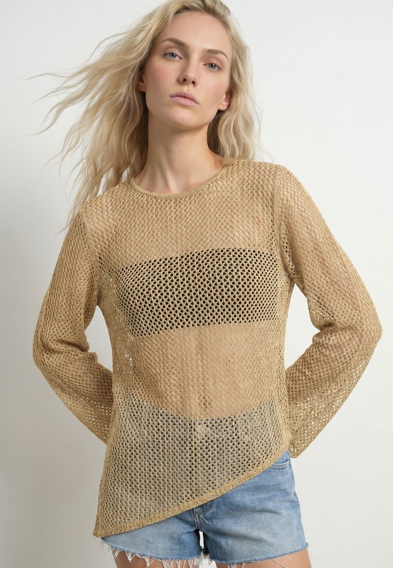 Femme aux longs cheveux blonds portant un pull en maille beige ajourée sur un top noir et un short en denim bleu clair, debout les mains dans le dos.