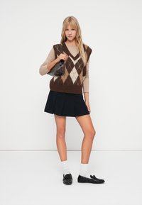 Brun og creme argyle strikket vest over en beige langærmet top, parret med en sort plisseret nederdel og sorte loafers. Lille skuldertaske holdt.