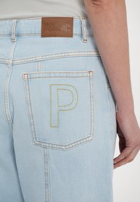 Jean en denim bleu clair avec des coutures orange et une lettre "P" jaune brodée sur la poche arrière. Comprend une étiquette de marque en cuir marron.