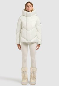 Witte pufferjack met een hoge kraag, voorzien van een chevronpatroon, gladde textuur en een logo op de mouw, gedragen over crèmekleurige leggings en laarzen.
