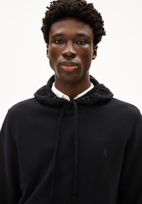 Zwarte hoodie met een zachte textuur, verstelbare koordjes en een subtiel logo op de borst. Gelaagd over een witte overhemd met kraag.