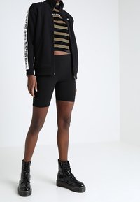 Persona con chaqueta negra de cremallera y texto blanco en las mangas, pantalones cortos de ciclismo negros, top recortado a rayas y botas de combate negras sobre un suelo blanco.