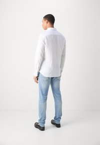 Mann in weißem Langarmhemd, hellblauen Jeans und schwarzen Loafers, der auf der linken Seite vor einem schlichten hellen Hintergrund steht.