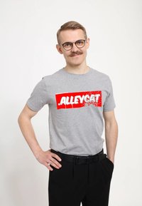 Šedé bavlněné tri�čko s velkým červeným grafickým prvkem a bílým nápisem "ALLEYCAT" v tučném, opotřebovaném stylu. Nosí se s černými kalhotami.