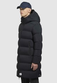 Cappotto lungo nero a puffer con cappuccio, caratterizzato da chiusura con zip e texture trapuntata orizzontale. Include tasche laterali e un'etichetta del marchio all'orlo.